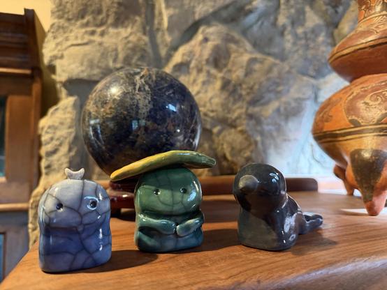 Three chunky little raku stoneware sculptures made by Laen Nash sit on a mantle near other sculptures. From left to right they are a dark sky moon nature spirit kodoma guardian whose color fades from smoky deep blue at the bottom to pale blue at the top with a little crescent moon on the head horns up; a changing seasons nature guardian who is very green and has a smile on its face with its little hands  joined together around its belly and a yellow-green leaf for a hat, and a magical raven kami nature spirit who is dark charcoal grey and looks like it might be up to something.