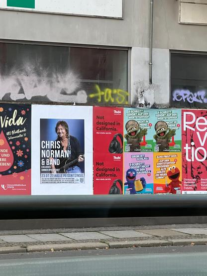 An urban wall displays several posters for concerts and two identical red Teufel posters like the ones in the original post with the text 'Not designed in California'. The wall has some graffiti and signs of wear.