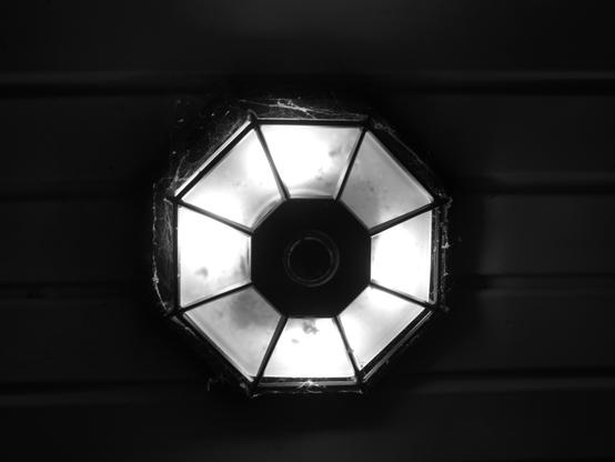 Looking up at a light fixture atop a porch which offers quadrilaterals. circles, and hexagons in it's assembly. There are cobwebs along it's edges illuminated by the light.