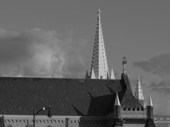 The rooftop and steeples of a church with geometric shapes including triangles, cones, and squares features in this photo set which is set against the sky containing a few clouds taken in morning light.