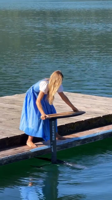 A woman in a dress launches herself from a dock onto a board elevated above the water and proceeds to pump her legs and so the board glides above the water surface