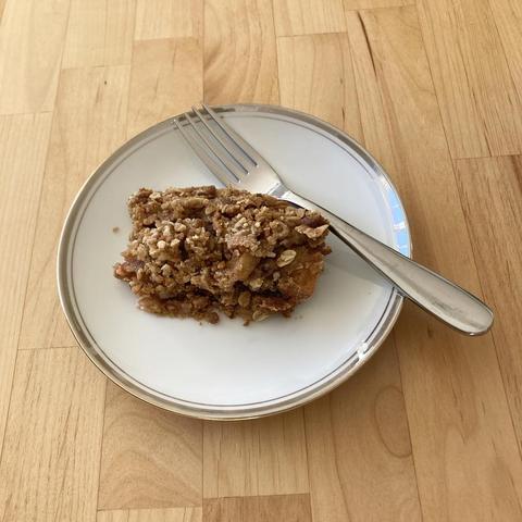 A serving of apple crisp on a white china plate with a thin gold stripe around the edge. A fork rests on the plate, which sits on a light wood tabletop.