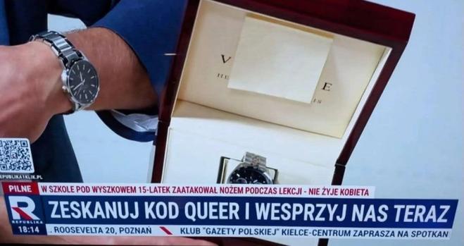 Kadr z TV Republika przedstawiający pudełko z zegarkiem, a na dalszym planie ów zegarek na ręce. W pasku jest napisane "Zeskanuj kod queer i wesprzyj nas teraz"