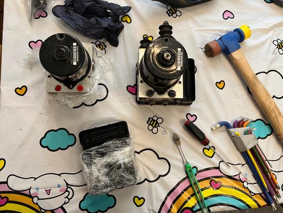 Parts for two ABS modules from a 2008 Volkswagen R32 on a Hello Kitty table cloth; my original, good pump module with red rubber hydraulic port plugs wrapped in plastic wrap to prevent particulates from getting into the other hydraulic channels. The new donor assembly is to its right. There's a hammer and some Torx keys which I used to do unspeakable things to free the halves of the donor module on account of a rusty bolt head.