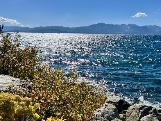 The sun reflects off a
lake. The shore is in the foreground. Mountains are in the background. In between them is Lake Tahoe.