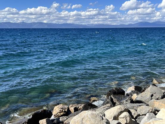 The shore of a lake. The lake is choppy. There are mountains beyond the lake and some clouds above the mountains.