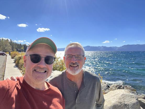 Two men on the shore of a lake on a sunny afternoon. The man on the left is wearing a baseball cap and sunglasses. The man on the right is wearing glasses. It is  57° F.