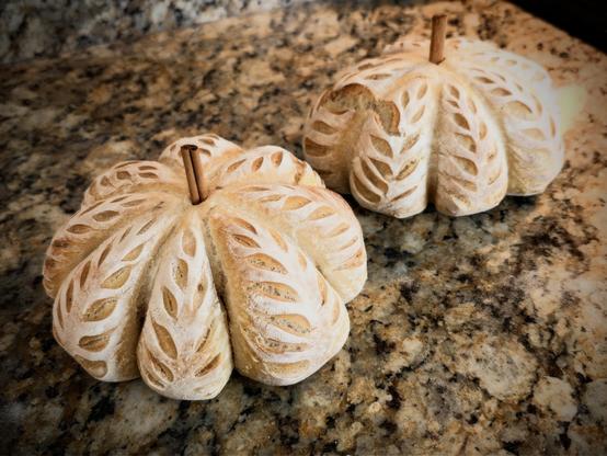 Two pale loaves shaped like pumpkins with leaf scores and cinnamon stick stems rest on a mottled granite countertop.