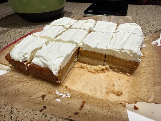 An 8 by 8 inch bake of pumpkin bars, cut into 12 pieces, with two missing. The bars are made of a bottom layer of shortbread, then pumpkin filling, and finally whipped cream, in roughly equal proportions.