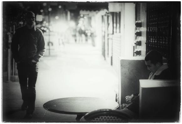North Beach. In monochrome. 8:07 p.m.

This is a picture about emptiness. Loneliness. The void. The infinite number of passing, unnoticed, near-encounters in our life, in space and time... moments that don't even register in our brains.

Two people on an urban sidewalk.

One strolls in the dark shadows on the left. He looks straight ahead. The other sits in the shadows at a cafe table on the right. He is looking down at his phone cradled in his hands. 

Between them, there is the glare and emptiness of a sidewalk... and in the far distance, the hint of a few people who will soon pass by and forget they were ever there.