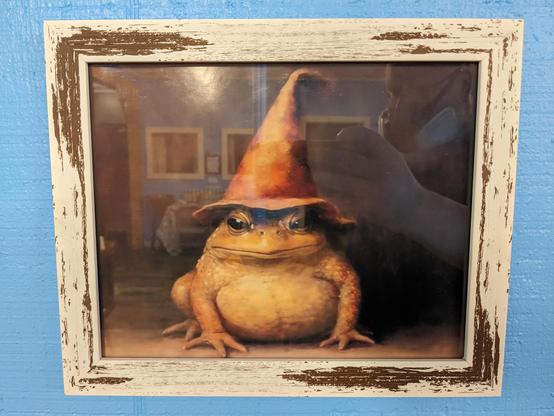 Painting of a yellow frog in a pointed orange hat with a floppy brim. It's in a frame with glass, with the frame intentionally scuffed on the top-left and bottom-right corners so as to look aged.

A bald man with glasses and wearing a 3M 6300 mask with N95 refillable cartridges can be made out in a reflection in the glass, holding a phone which is evidently taking the photo of the painting.

In the background, also caught in the reflection, we see a wall opposite this one with other framed paintings.