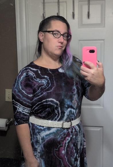 Mirror selfie of a woman with purple and blue hair, shaved on one side, wearing glasses and a dark purple, blue, and white tie dyed dress cinched at the waist with a wide white eyelet belt.