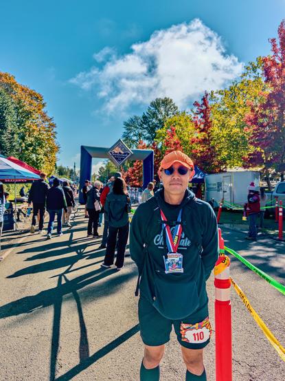 Me at the finish area in the sun after the race, in an orange cap and sunglasses, wearing the finisher medal and bib 110, the blue Snohomish Running Co arch and red autumn trees behind me.
