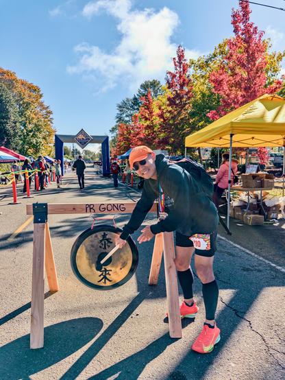 Me hitting the PR GONG with a mallet at the finish area, in orange shoes and black compression socks, under a clear blue sky.