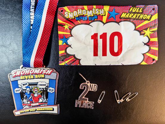 The finisher medal, bib 110 for the full marathon, a 2nd place charm and four safety pins on a black surface.