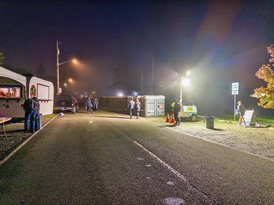 The race start area before dawn in thick fog: a food truck, portable toilets and a light tower glowing over a dark road, a few runners walking in.