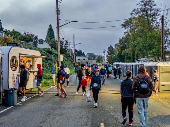Runners in warm layers walking past a coffee truck and a long row of portable toilets under a grey sky before the start.