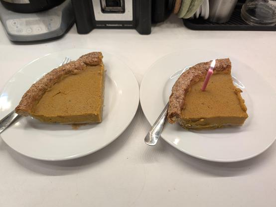 Two generous slices of pumpkin pie on white plates, one with a lit pink birthday candle in it.