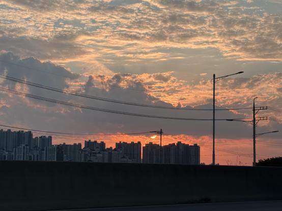 A sky with partial cloud cover. Bits of blue shine through toward the top of the frame, and the bottom half is hues of orange. The sun peeks out just below a heavy cloud formation on a trajectory to set behind a high rise apartment complex. The found has a few light poles and wires. The lower quarter is a dimly lit concrete barrier.