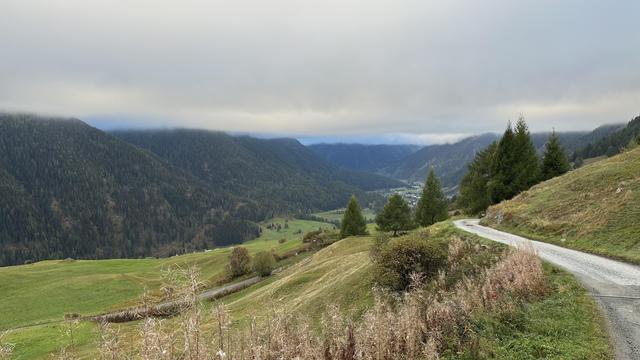 View past pastured hillsides towards the lower Engadine valley in the direction of Lavin. A low cloud ceiling obscures all the peaks but patches of blue are visible on the horizon.