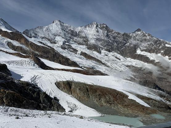 From Felskinn, a crevassed glacier flows down in several tongues between brown rock bands, beneath three snow-covered peaks. A glacial lake sits in the bottom right. Lift cables cross the frame and wisps of cloud cling to the slope.