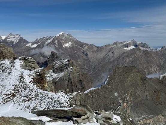 Panorama from Mittelallalin: fresh snow on rocks and a ridge in the foreground. Beyond a deep valley, a range of grey-brown peaks lightly dusted with snow, with clouds clinging to their flanks.