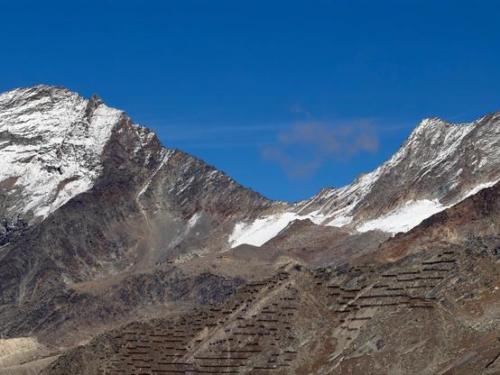 Two rocky ridges dusted with fresh snow form a V-shaped col in the centre, against an intense blue sky. Station buildings sit at the foot of the col, and neat rows of avalanche barriers line the slope in the foreground.