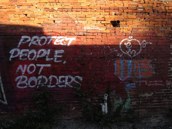 Eine stark verwitterte Backsteinmauer, deren Fugen dringend ausgebessert werden müssten; darauf hat jemand mit Sprühfarbe den Schriftzug „PROTECT PEOPLE, NOT BORDERS“ hinterlassen. Sonnenlicht fällt auf das obere Drittel der Mauer, während der Rest im Schatten liegt. Es sind noch weitere Graffiti zu sehen – vermutlich das „Tag“ (die Signatur) einer Person. Aus dem Mauerwerk ragen einige Rohre hervor. Das Foto entstand in Pittsburgh, Pennsylvania.

A heavily weathered brick wall with mortar joints in urgent need of repair; someone has spray-painted the words "PROTECT PEOPLE, NOT BORDERS" on it. Sunlight falls on the upper third of the wall, while the rest remains in shadow. Other graffiti is also visible—presumably a person's "tag" (signature). Several pipes protrude from the masonry. The photo was taken in Pittsburgh, Pennsylvania.