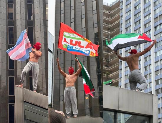 A protester in Brazil stands upon a rooftop. He only wears grey sweats and a red scarf as a mask to hide his face. He's well fit. Three pictures of him with different flags: trans pride, in support of Lula and the Palestinian flag.