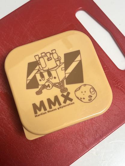 A small square Tupperware with an image of a cartoon version of the MMX spacecraft.