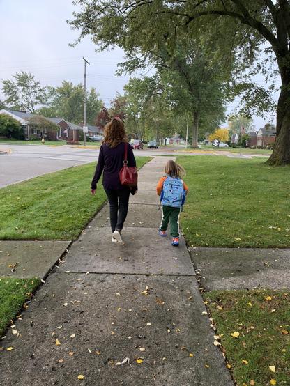 A woman and a young child walk away from the camera down a neighborhood sidewalk on an overcast autumn day. The woman has curly hair and is wearing a dark long-sleeved shirt, dark pants, and carrying a red shoulder bag. The child is wearing an orange shirt, green pants with a white stripe, and a blue backpack with a pattern on it. Fallen yellow leaves are scattered across the sidewalk and grass, and mature trees line the street.