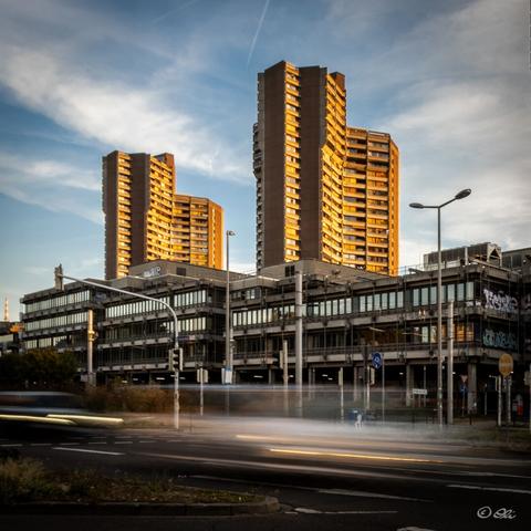 Two (of the three) "Neckaruferbebauung" towers glow  in the evening light above lower office building with graffiti. Cars on the road in front are blurred by a long exposure. Blue sky with clouds.