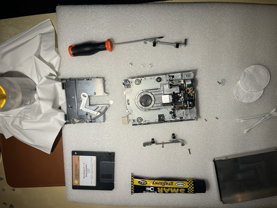 Partly disassemble internal floppy diskette drive from an Amiga 500 computer, shiny metal and electronics parts placed on a soft plastic pad, some graphite grease, isopropanol, cosmetic pads and ear buds are also in field of view