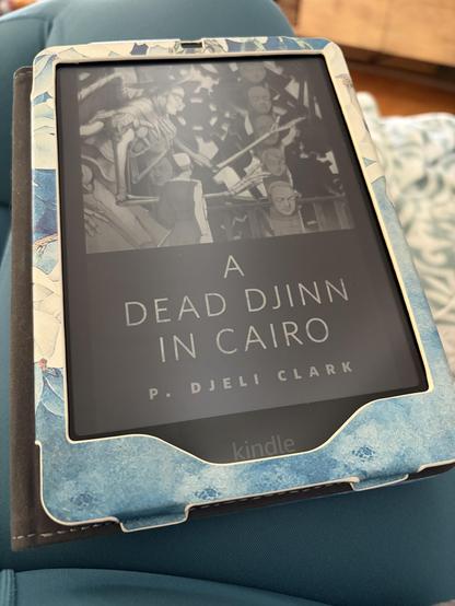 A photo of a kindle on Ashe’s teal-clad lap. The cover on the device is for A Dead Djinn in Cairo by P. Djeli Clark.