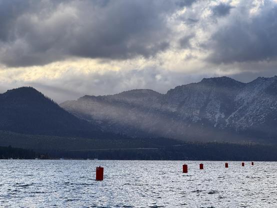 Mountains and clouds with light rays spilling between them illuminating a lake. Red channel markers are visible in the water.