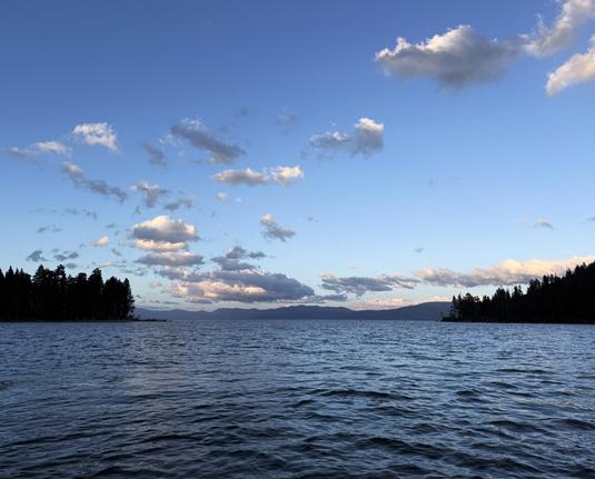 The opening of a bay into a large lake. Pine trees cover either side of the land and several small clouds are lit with late evening light.