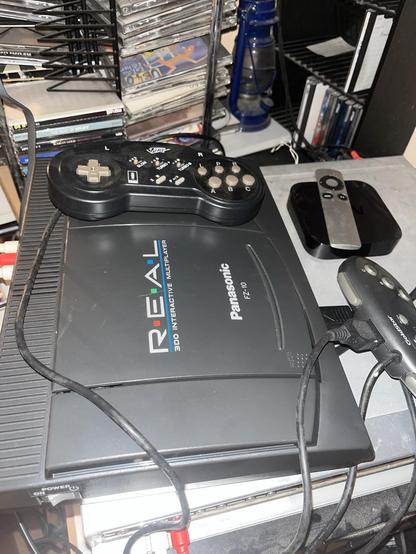 An image of a Panasonic 3DO FZ-10 gaming console with controllers on a cluttered surface. In the background, there are stacks of CDs and a small media player.