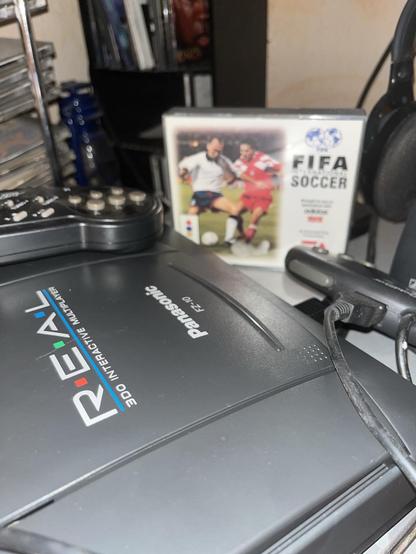 A Panasonic 3DO gaming console is prominently displayed, with a remote control and a FIFA soccer video game case next to it. In the background, there are shelves filled with DVDs or CDs.
