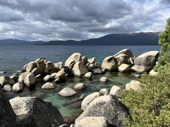 Large granite boulders at a lake’s edge. Mountains behind the far shore of a lake. The sky is mostly overcast, but the sun shines on the granite boulders in the foreground.