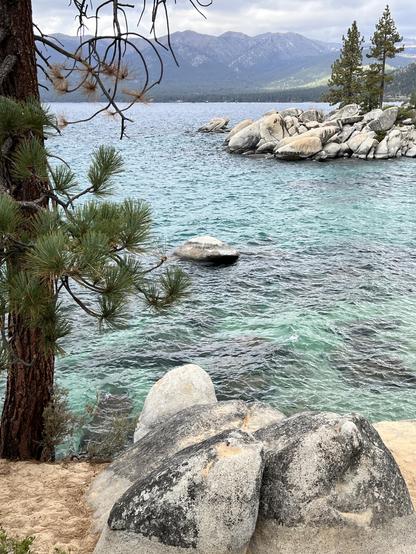 Lakeside scene: grante boulder in the foreground, the lake in the middle distance, and mountains in the background. The sky is overcast.