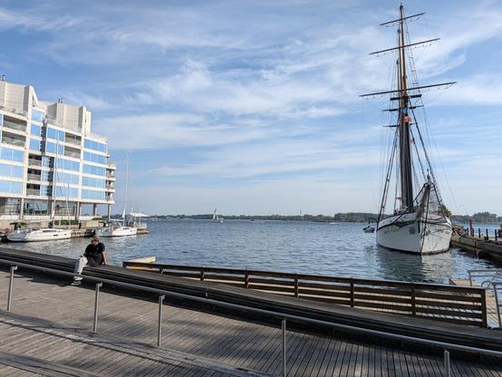The same blue-white sky now over a view of Lake Ontario from a boardwalk, with a multistory glass and concrete building to the left of the frame and a white boat with a tall mast and no sails moored to a dock on the right.