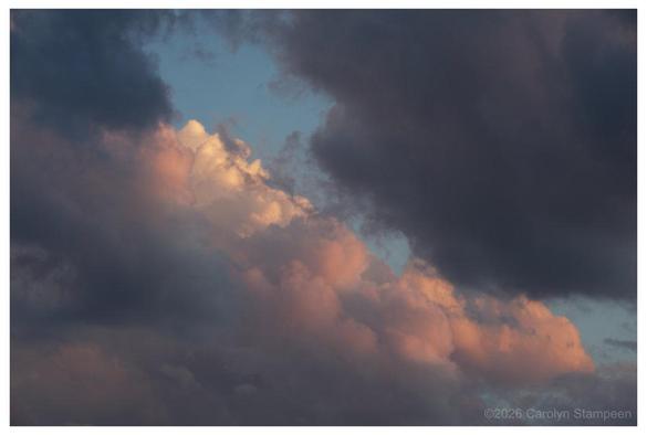Pink, orange, and white fluffy clouds are behind dark purplish-grey that are visible in the top right and left corners of the image.  Behind them, we can see glimpses of blue sky.