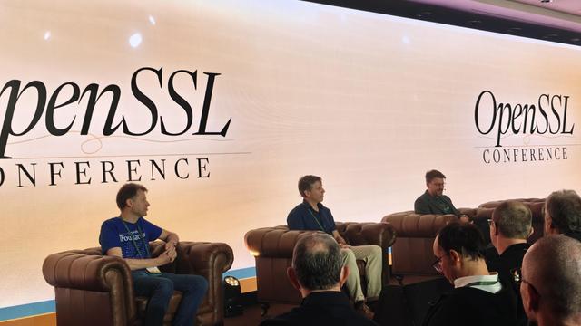Three white dudes on stage sitting in comfy arm chairs, in front of a huge video wall.