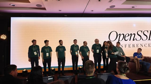 8 people in green branded OpenSSL T-Shirts standing on the left of a stage in front of a video wall.