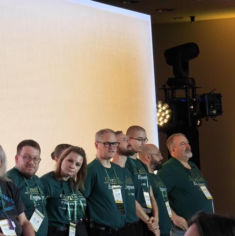 Another 8 people on the right end of the same stage, in front of a video wall, wearing the same green OpenSSL-branded shirts.