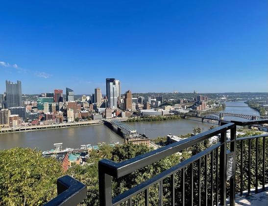 View of a city from above and across a curving river. Several bridge crossing the river are visible, and tall buildings interspersed with lower ones. The sky is bright clear and blue.