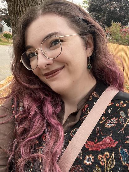 Tulip smiling, wearing dark plummy-brown lipstick, a floral brocade vest, and silver dangly earrings. there’s some reddish trees in the distance