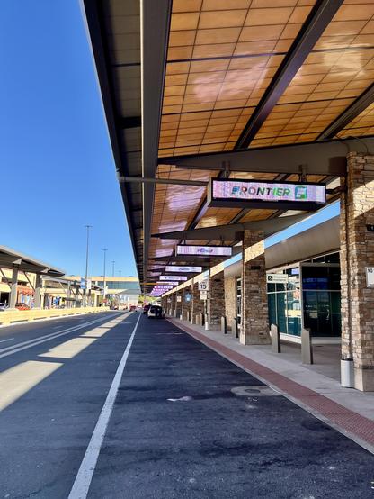 Looking down the curb outside an airport. Roof on the top of the image, pavement on the bottom. It is a sunny day. The lines of the image seem to converge in the distance to a point just left of and below the center of the image.
