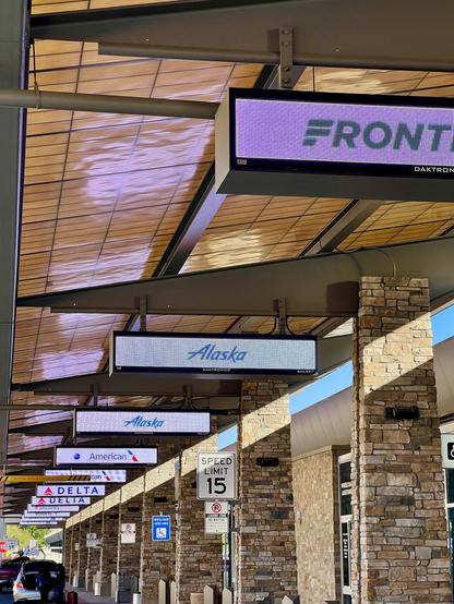 Curbside at an airport. Signs indicating different airlines hang from the ceiling. The scene is outdoors.