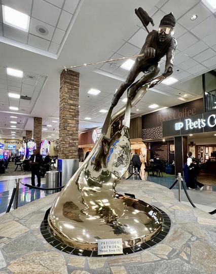 A large sculpture of a dowhill ski racer rises from a stone floor. The statue’s base is a shiny brass. The scene is inside an airport.